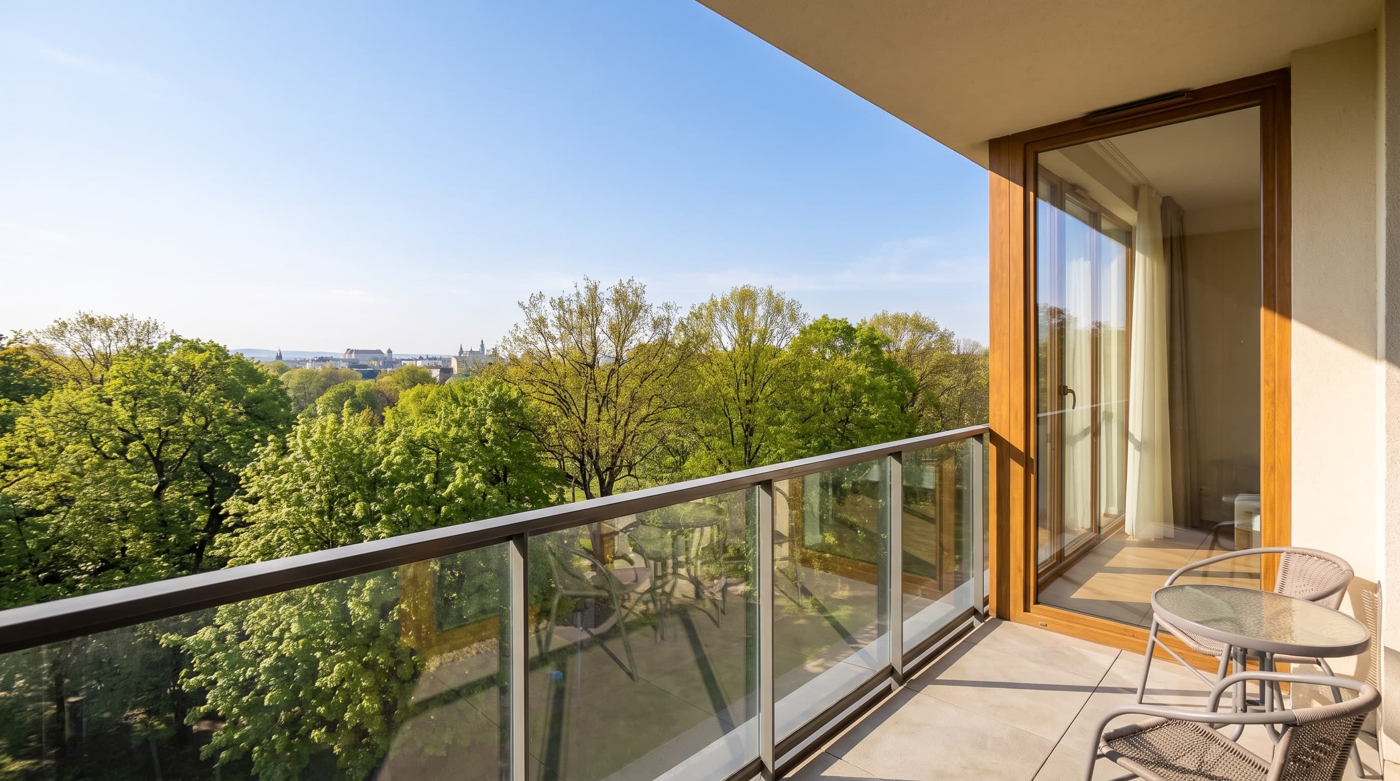 The balcony of A21 with two chairs, a small table and planters behind a glass balustrade, the park canopy below and the Kraków skyline on the horizon